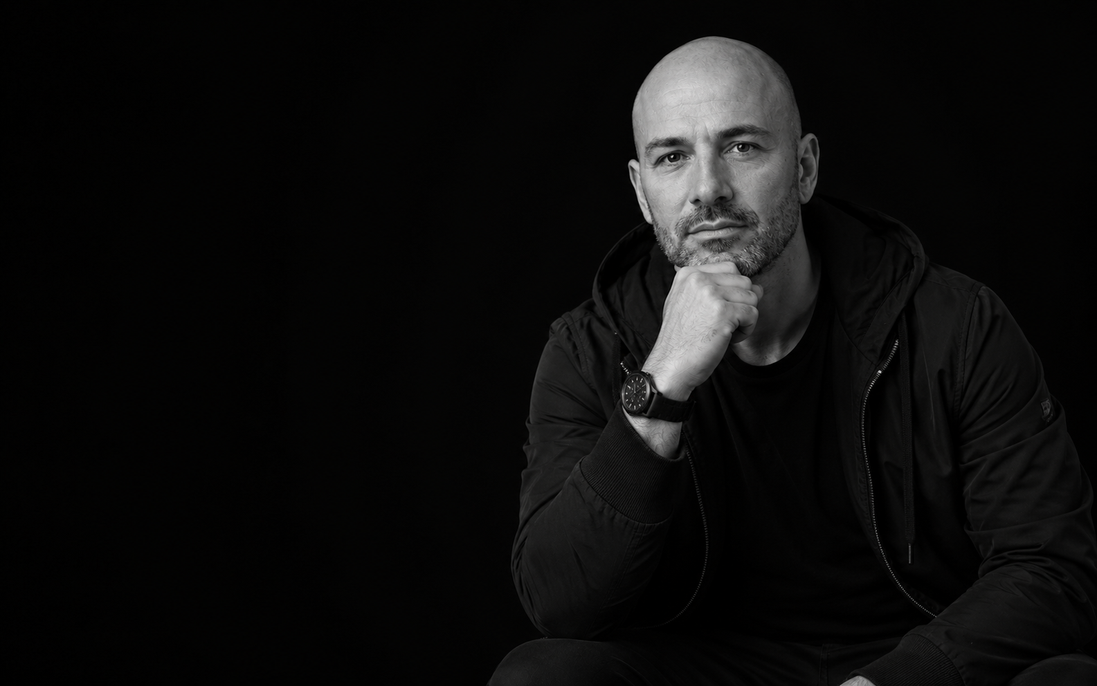 Bald man in a black jacket, hand on chin, confident portrait
